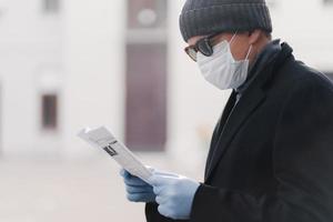 Cropped shot of man wears facial medical mask and protective rubber gloves, prevents coronavirus infection, reads newspaper, poses outdoor against blurred background. Quarantine, Covid-19 concept photo