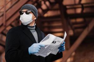 Virus spread and quarantine concept. Serious man wears sunglasses, hygienic mask and rubber gloves, looks away, reads press, protects himself from risk of viral disease or coronavirus, air pollution