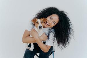 Happy curly woman tilts head, holds pedigree dog, has cheerful expression, smiles pleasantly, has curly hairstyle, wears white t shirt, isolated. Afro lady petting favourite domestic animal. photo