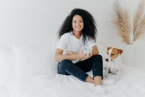 Glad dark skinned Afro American female feels relaxed, poses in bedroom on comfortable bed with pedigree dog, drinks hot beverage, has morning coffee, smiles happily, enjoys spare time at home. photo