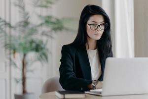 Young serious businesswoman works with laptop computer at workplace in office, wears optical glasses and formal costume, concentrated at screen. Job, occupation, business development concept photo