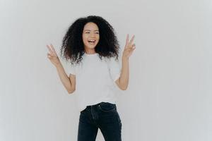 Carefree woman with Afro hairstyle raises hands up, shows victory gesture or peace sign, looks gladfully aside, dressed in casual wear, smiles relaxed, sends hello, isolated over white background. photo