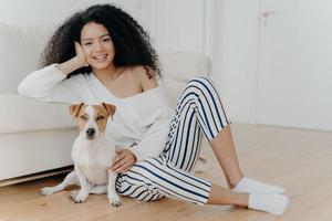 Horizontal shot of beautiful curly woman leans at sofa, sits on floor near pedigree dog, wears sweater, striped pants and socks. African American lady poses with pet. People and animals concept photo