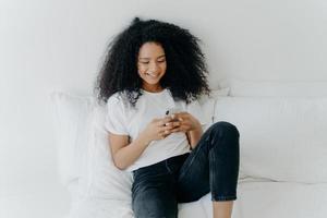 una mujer afro hermosa y relajada con expresión alegre usa un teléfono móvil moderno, navega por Internet, posa en una cama blanca y cómoda, está conectada a wifi, navega por la página web, disfruta de un ambiente doméstico foto