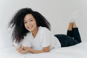 Horizontal shot of curly ethnic woman with pleasant smile, lies on comfortable bed, raises legs, uses modern cellphone, reads text message, enjoys high speed internet connection. People and bed time photo