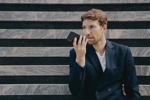 Male executive worker dressed formally holds smartphone near mouth talks on speakerphone photo