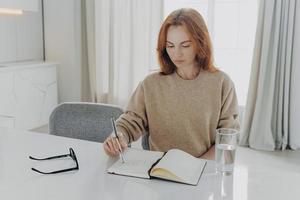 serious redhead young woman makes notes in notepad sits at table photo