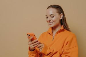 Happy charming young woman holding smartphone in hands and smiling, chatting in social networks photo