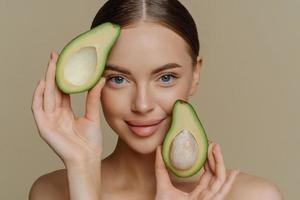 Close up shot of tender blue eyed woman looks directly at camera holds halves of avocado near face chooses healthy nutrition has perfect smooth skin isolated over beige background. beauty concept photo