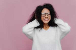 Portrait of joyous pleasant looking African American woman covers both ears with hands, hears loud music, smiles broadly, wears spectacles and white warm sweater, poses indoor, being very emotional photo