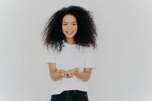 Glad dark skinned woman has cupped hands, asks to give something, smiles plesantly, has bushy frizzy hair, wears t shirt and jeans, isolated over white background. People and charity concept photo