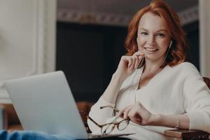 Positive redhead woman has video call with friend via laptop computer and earphones, enjoys time for chatting via web camera, has online web seminar during remote job, poses in coworking space photo