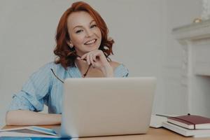 Self employed female freelancer has gentle smile on face, works on startup project, poses with laptop computer at desktop, searches information for productive work, sits indoors, holds eyewear photo