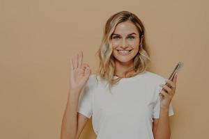 Happy cheerful female holding smartphone and showing okay gesture, being happy after pleasant conversation photo