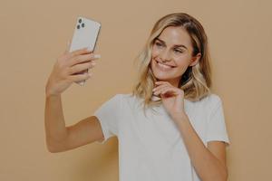 Cute lovely female with blonde hair making selfie on modern mobile phone and smiling, taking picture of herself photo