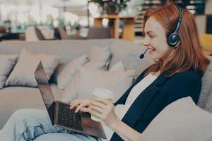 Smiling young female teacher with wireless headphones and laptop having video conference in cafe photo
