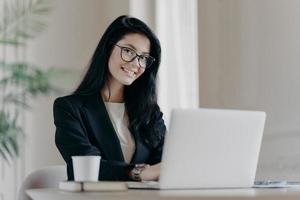 una mujer positiva de cabello oscuro investiga, busca información en una computadora portátil, se conecta a Internet inalámbrica, es feliz, usa ropa formal y anteojos bebe café. economista trabaja en linea foto