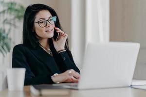 Confident serious business lady has phone conversation, poses in front of laptop computer, wears optical glasses for vision correction, poses at work place, makes reserach work, dressed formally photo