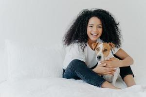 People, pet care and friendship concept. Smiling Afro woman with glad expression cuddles pedigree dog, sit on comfortable bed, copy space area on white background, going to sleep or have rest photo