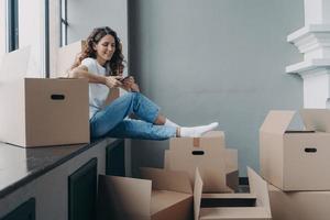 Cheerful european woman is sitting on windowsill packing boxes and chatting on smartphone. photo