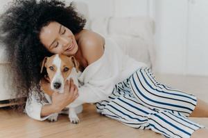 Affectionate young woman hugs dog with love and care, keeps eyes closed from pleasure, smiles gently, has healthy dark skin, poses on floor, petting animal. People, friendship and pets concept photo
