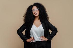 Half length shot of happy Afro woman dressed in elegant black clothes, keeps both hands on hips, wears optical glasses, smiles broadly, isolated over brown background. Female director in good mood photo