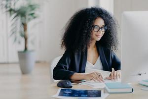 Freelance Afro woman works remotely, writes information, focused at computer screen with delighted expression, poses at workplace in spacious cabinet, potted plant in background. E learning concept photo