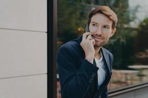 apuesto hombre de negocios profesional urbano escuchando al cliente en el teléfono celular mientras está parado afuera foto