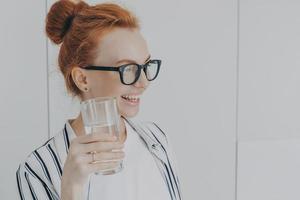 Thirsty woman with red hair combed in bun drinks still water prevents dehydration holds glass photo