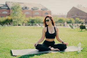 Relaxed brunette woman sits on fitness mat on lotus yoga pose wears sunglasses cropped top and leggings looks aside drinks water after training breathes deeply fresh air. Healthy lifestyle concept photo