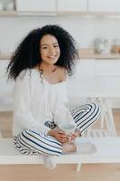 Image of lovely curly haired lady drinks coffee or tea from white mug, wears fashionable white sweater, striped pants, poses at kitchen against blurred background. People and lifestyle concept photo