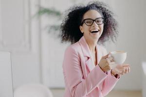 Overjoyed curly haired woman laughs happily while drinks hot coffee or latte, wears transparent spectacles, has fun during break in office, shows white teeth, being professional entrepreneur photo