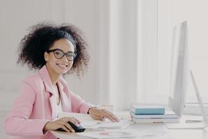 Prosperous curly female entrepreneur checks documents, studies contract, works on computer, wears glasses and pink jacket, poses at workplace, smiles broadly, happy almost finishing work in time photo