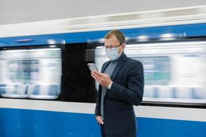 Young man manager uses cellphone, prevents spreading of Coronavirus, poses against subway train, poses at platform, checks news online, wears medical mask. Epidemic situation, health care concept photo