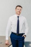 Vertical shot of handsome unshaven cheerful man wears formal clothes, keeps hand in pocket, stand near office table, has positive smile. Prosperous businessman being at work place. Business concept photo