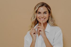 Young beautiful positive female with blond dyed hair touching lips with finger and smiling at camera photo