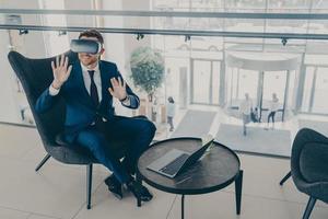 Smiling office worker sitting in company center lobby with laptop and VR headset goggles photo