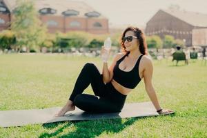 Happy young sporty European woman has rest after training poses on fitness mat with bare feet drinks water dressed in activewear sunglasses leads healthy lifestyle. People workout hydration concept photo
