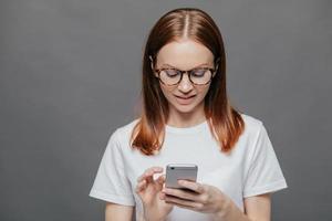 Portrait of pleasant looking relaxed woman holds modern cell phone, types text message on cell phone, connected to wirelesss internet, dressed in white t shirt, isolated over grey wall. Technology photo