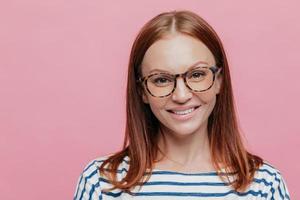 Close up shot of good looking brown haired woman with no make up, wears optical glasses, has gentle smile on face, dressed in striped clothes, models over pink background. Positive emotions concept