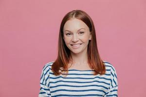 Headshot of pretty smiling European woman with charming smile, wears striped jumper, has brown hair, looks directly at camera, isolated over pink studio background. People, joy, happiness concept photo