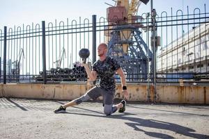 Young bearded male athlete training in industrial zone in sunny day, kettlebells exercises outdoors, urban background photo