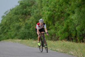 Bicycle racer in helmet and sportswear training alone on empty country road, fields and trees background photo