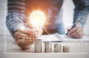 Man hand holding light bulb on coins stack on the wooden table , Saving ideas and investment budget, Creative ideas concept, Stock market or forex trading graph and candlestick chart suitable photo
