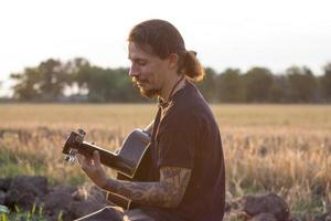 Young tattooed male play on acoustic guitar in summer fields during beautiful sunset photo