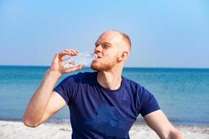 joven atleta masculino bebiendo agua dulce de una botella de plástico en la playa foto