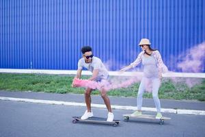 pareja joven montando en patinetas con bomba de humo de color, niño y niña con ropa informal pasando un buen rato, colores rosa y azul foto