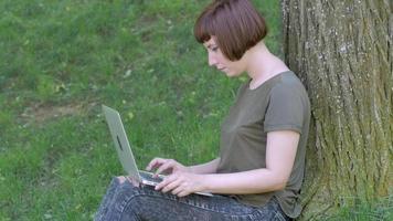 una joven trabaja con una laptop al aire libre en el parque de verano, una mujer con una computadora en la hierba verde 4k tiro video