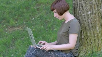 Young woman work with laptop outdoors in the summer park, female with computer on the green grass 4k shot video