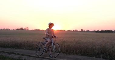 jovem mulher com chapéu andar de bicicleta em campos de trigo de verão